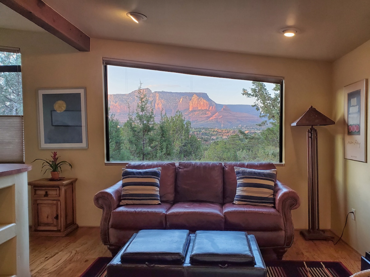 A spacious living area features a brown leather sofa adorned with striped throw pillows. A large window showcases vibrant red rock formations bathed in sunset light, surrounded by lush greenery. A wooden coffee table sits in front, adding to the cozy atmosphere.