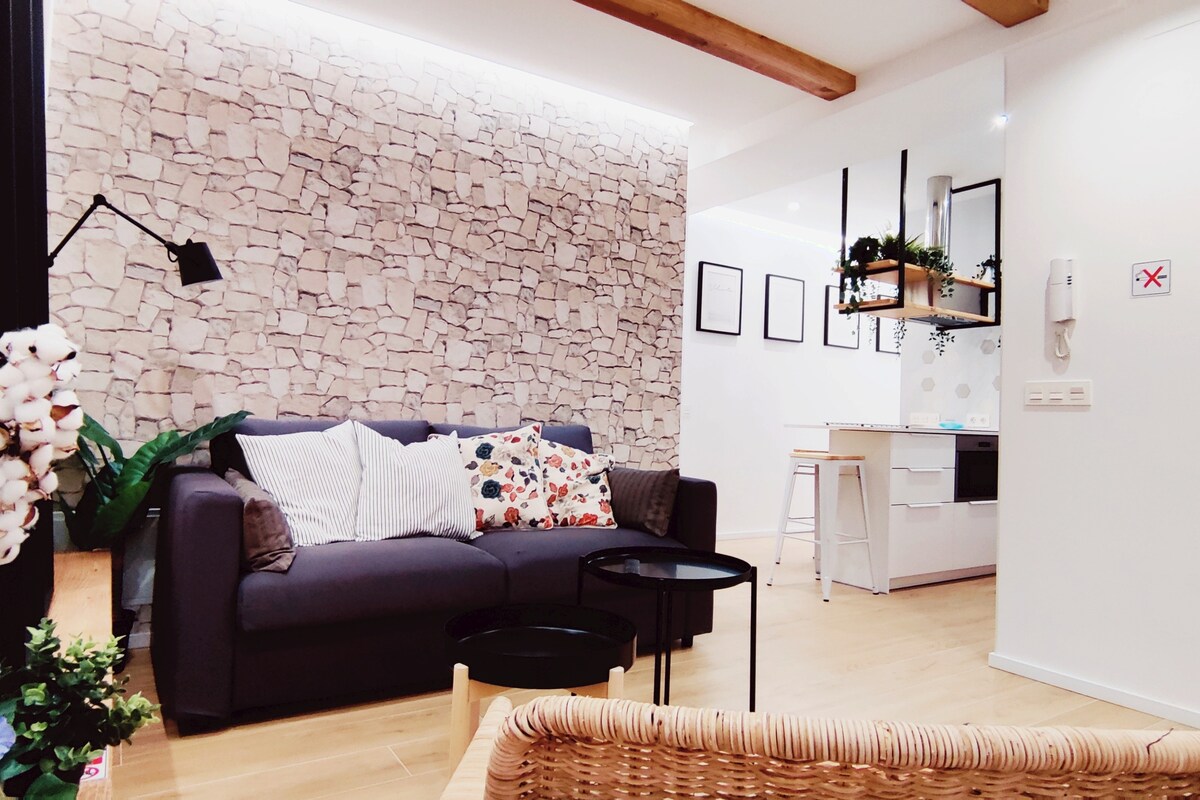 A modern living area features a stylish gray sofa adorned with patterned cushions. Two small black coffee tables are positioned in front. A textured stone accent wall adds character, while light wood flooring enhances the space's warmth. The kitchen is subtly visible in the background.