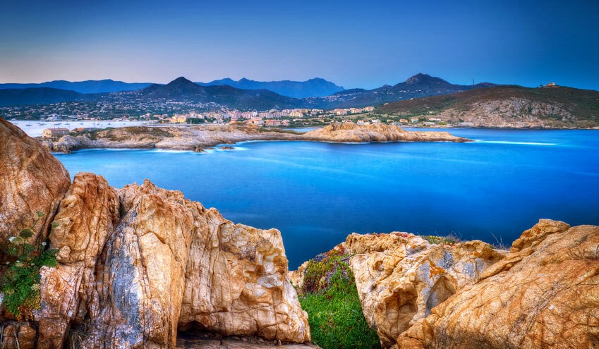 A panoramic view of the calm blue sea is framed by rugged, golden rocks in the foreground. The shoreline features gentle waves, while distant mountains create a serene backdrop against the evening sky, which transitions from deep blue to soft hues.