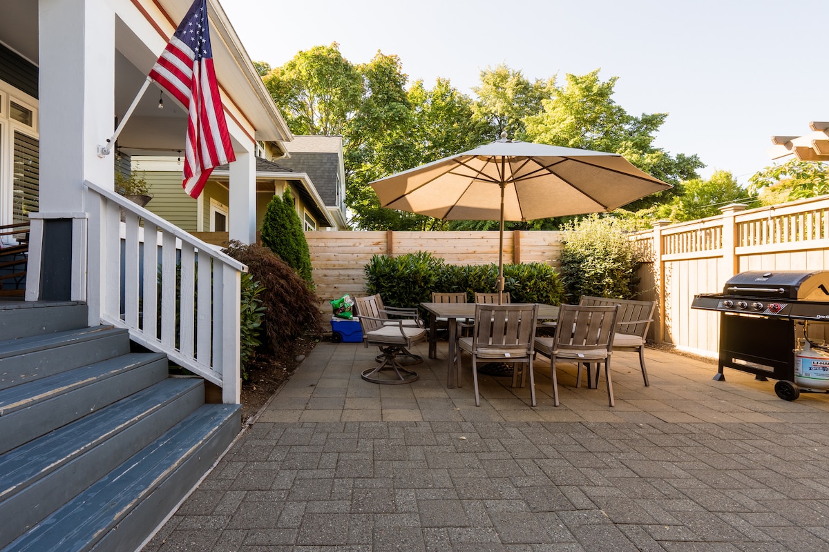 An outdoor patio area is highlighted, featuring a large umbrella over a round table surrounded by several chairs. A grill is positioned nearby, and a flag is displayed on the house. Lush greenery provides a natural backdrop, creating a serene outdoor space.