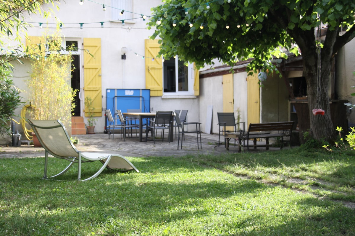 A serene outdoor space is showcased, featuring a grassy area with a lounge chair and several seating arrangements. Soft lighting strings are draped overhead, while yellow shutters frame the background, enhancing the garden's welcoming ambiance.