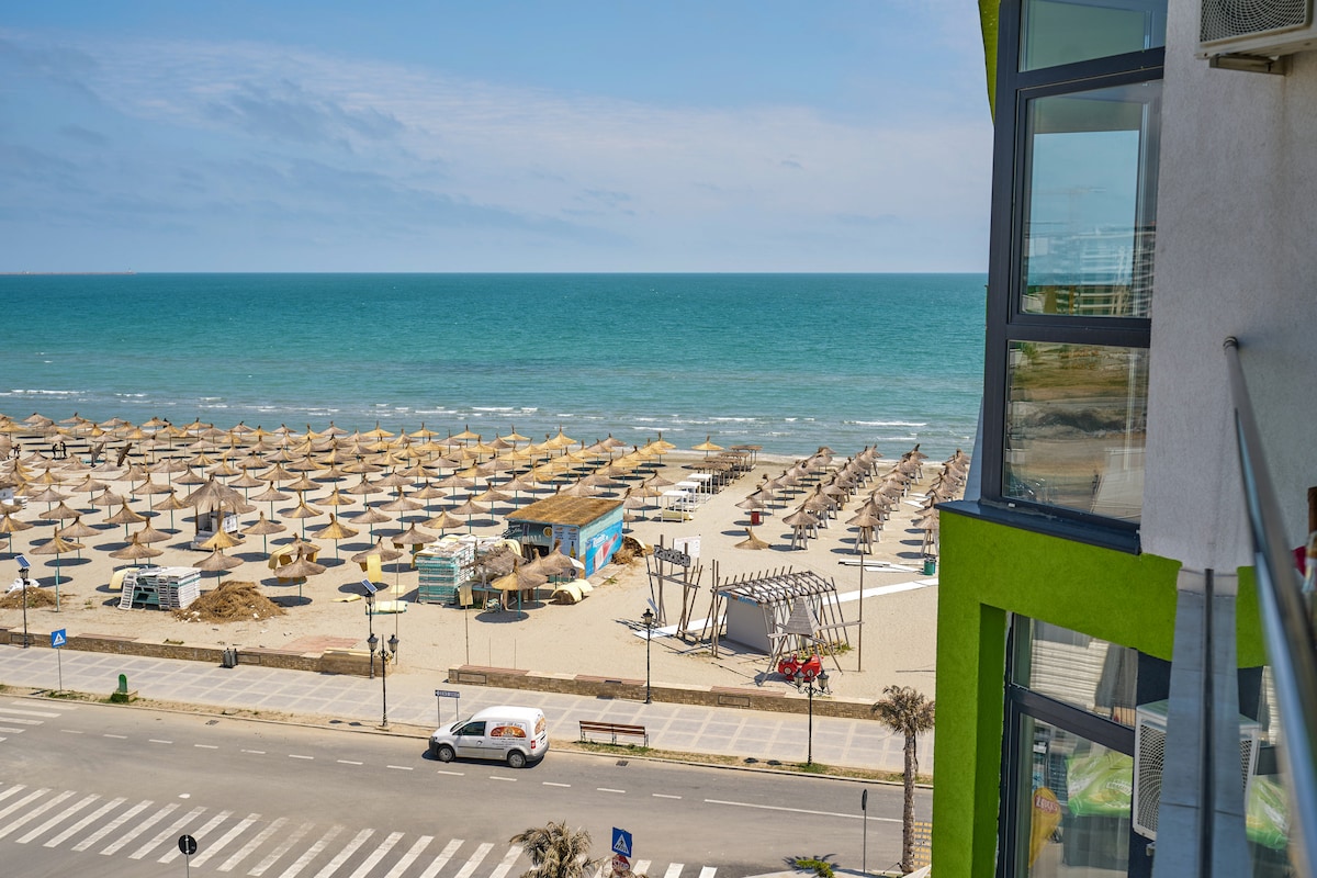 A panoramic view of the beach is displayed, featuring numerous sun loungers and umbrellas arranged across the sandy shore. The calm sea extends to the horizon, with gentle waves lapping at the beach. A clear sky complements the seaside scene, enhancing the tranquil ambiance.