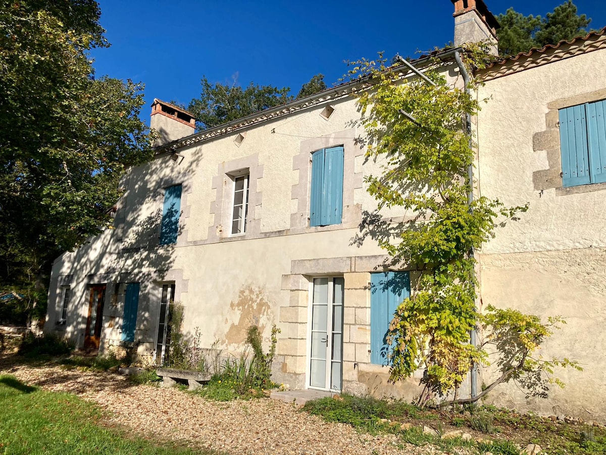 The charming stone house features light blue window shutters and a rustic facade. A modest garden area with gravel and greenery is visible in the foreground. Sunlight highlights the natural elements, providing a warm ambiance to the surrounding landscape.