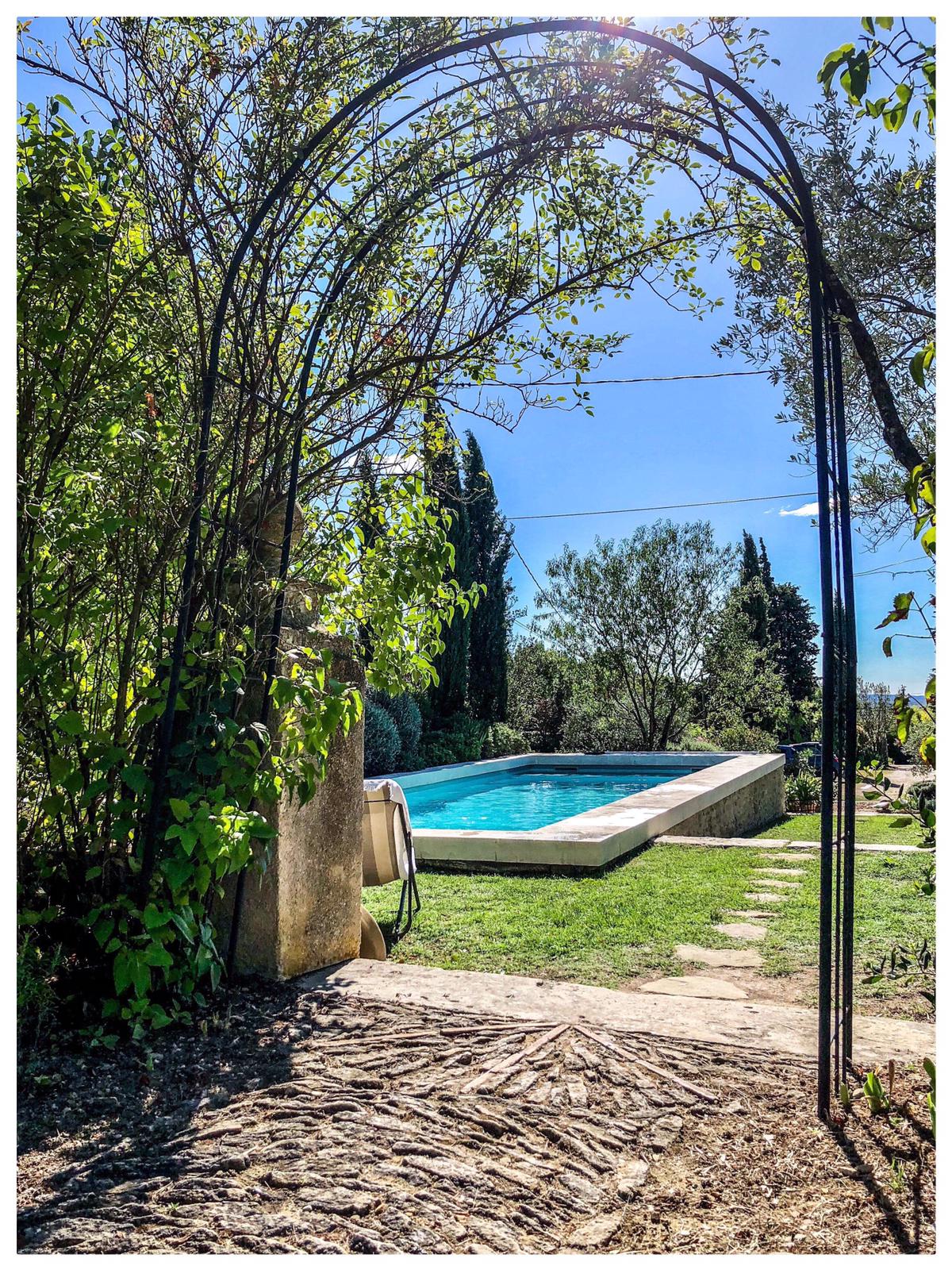 An archway covered in greenery frames a view of a swimming pool surrounded by lush landscaping. The clear blue water of the pool reflects sunlight, and tall trees can be seen in the background, contributing to a serene outdoor setting.
