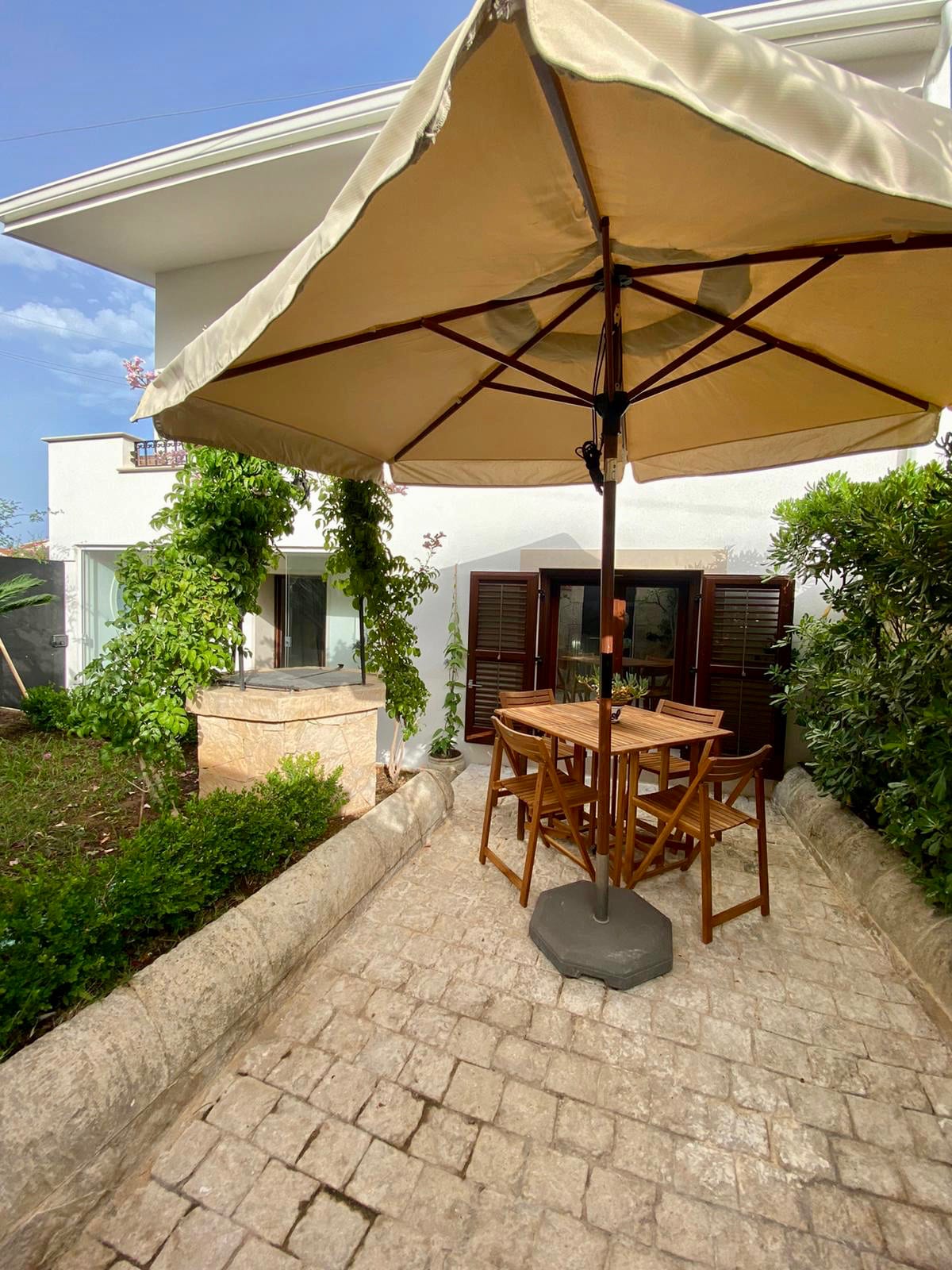 A small private garden area features a wooden table and four folding chairs, shaded by a large umbrella. Lush green plants frame the space, while partially opened wooden shutters allow natural light into the nearby living area.