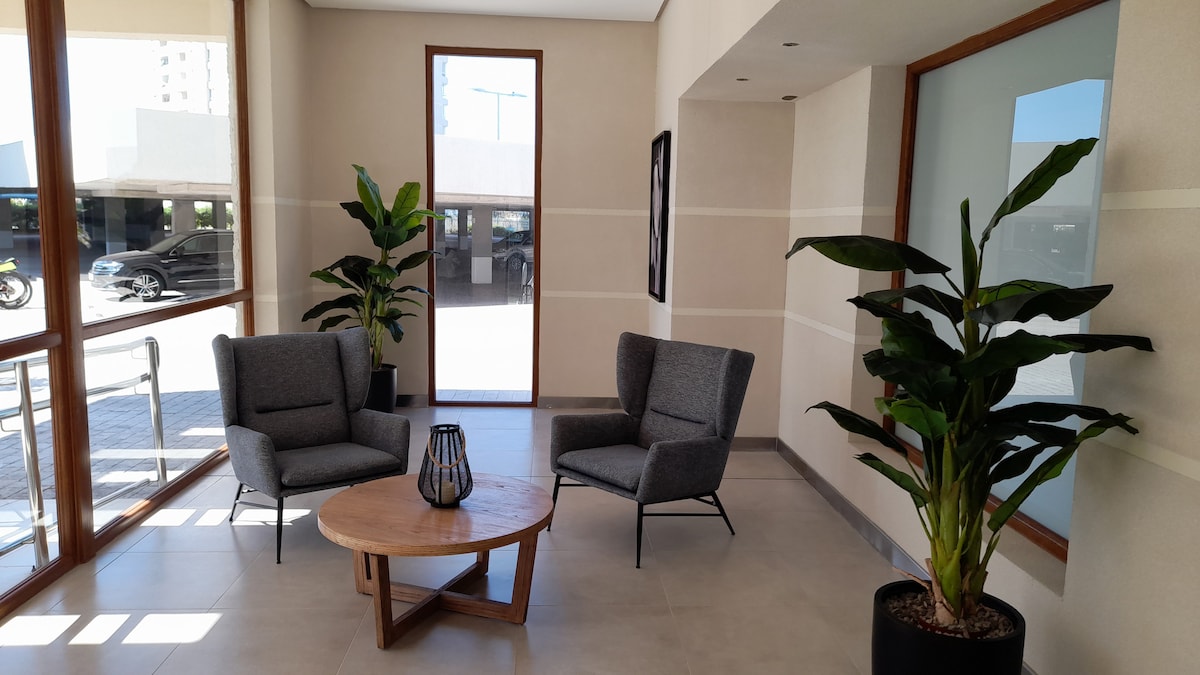 A welcoming lobby area is featured, showcasing two comfortable gray armchairs positioned around a wooden coffee table. Lush green plants are placed in modern pots, complementing the natural light streaming in through large glass windows, which offer a view of the outdoor space.