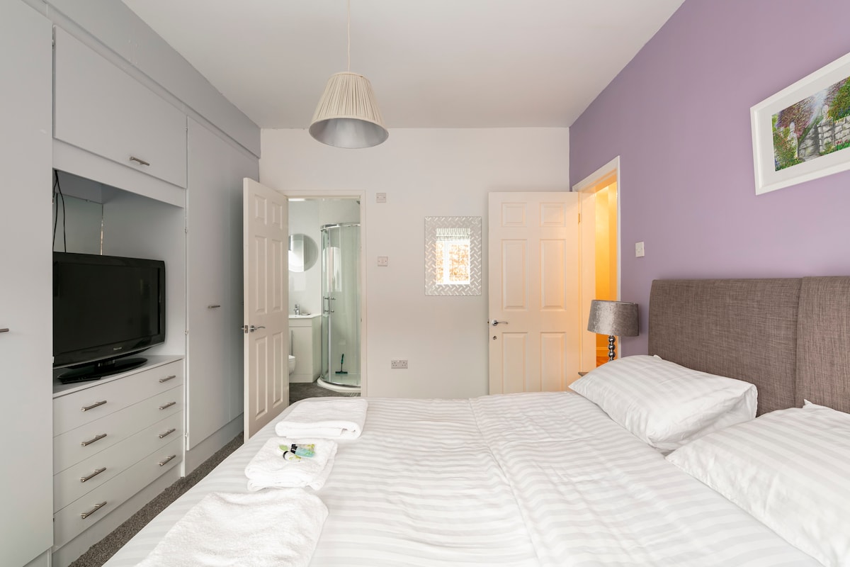 A guest room is presented with a zip link bed, capable of accommodating a superking or twin configuration. A television is positioned across from the bed, while an ensuite bathroom is visible through an open door. Soft towels are neatly arranged on the bed.