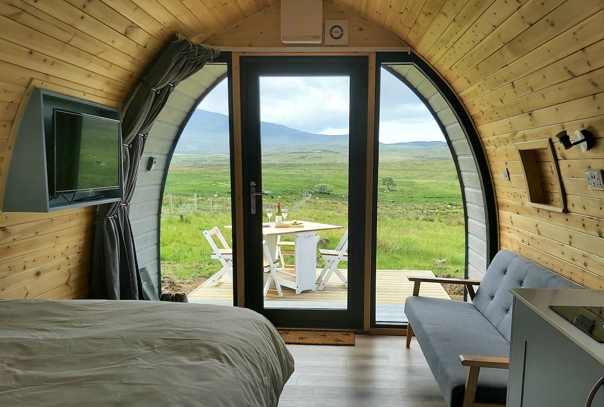 The interior features wooden walls and a curved ceiling with a large glass door leading to the outdoor deck. A cozy sofa and dining table are visible, alongside a television mounted on the wall. Spacious views of the surrounding landscape are presented through the door.