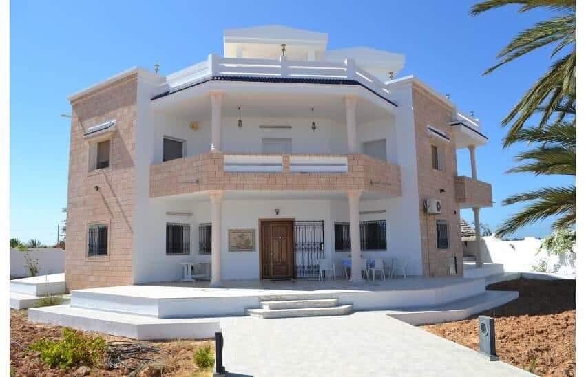 The three-story villa features a light-colored stone exterior with multiple balconies and large windows. A wide staircase leads to the entrance, and outdoor seating is visible in front. Lush greenery surrounds the property, contributing to a serene and inviting environment.