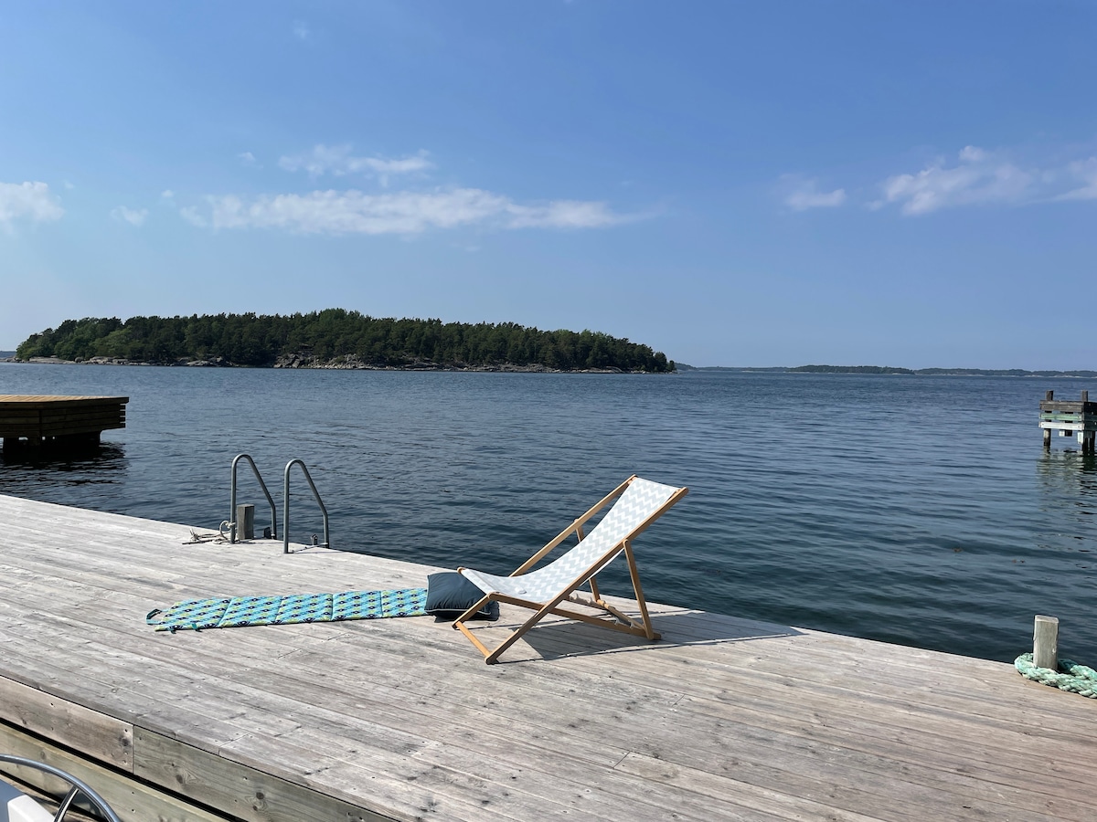 A wooden dock extends over calm water, featuring a sun lounger positioned for relaxation. In the distance, an island is visible, surrounded by greenery. A swimming ladder is attached to the dock for easy access to the water.