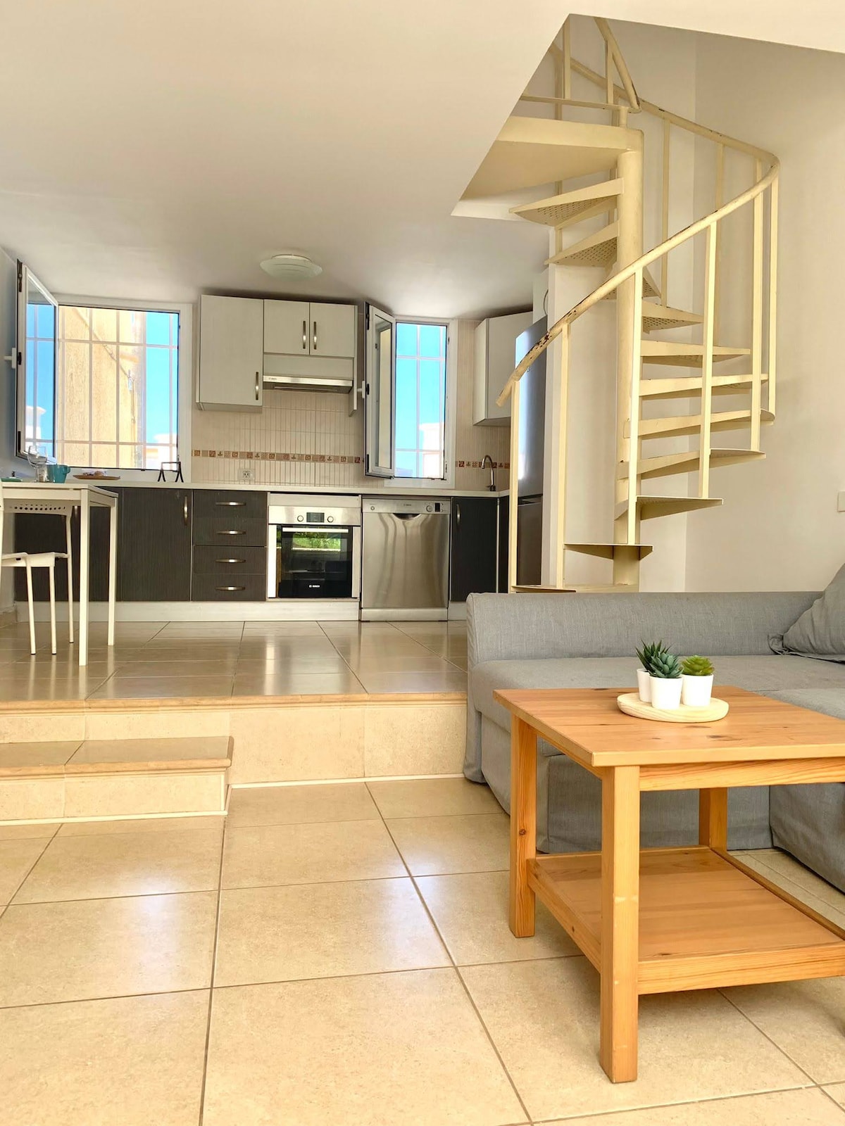 The living area features a gray sofa and a wooden coffee table with a small plant. A spiral staircase leads to the upper floor. The kitchen is visible in the background, equipped with modern appliances and flanked by large windows that provide ample natural light.