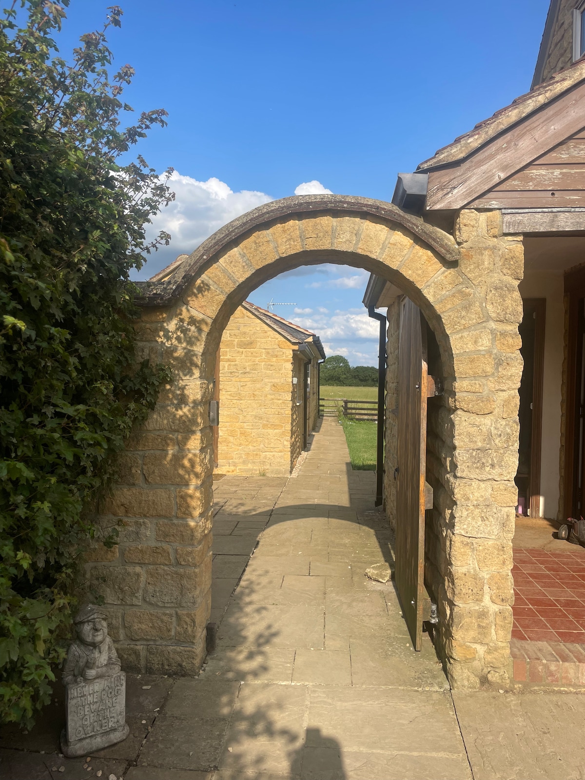 A stone archway frames a pathway leading to the annex, bordered by lush greenery. Sunlight casts a warm glow on the textured stone walls, while a clear blue sky with fluffy clouds extends in the background. A sense of openness to the countryside is visible.