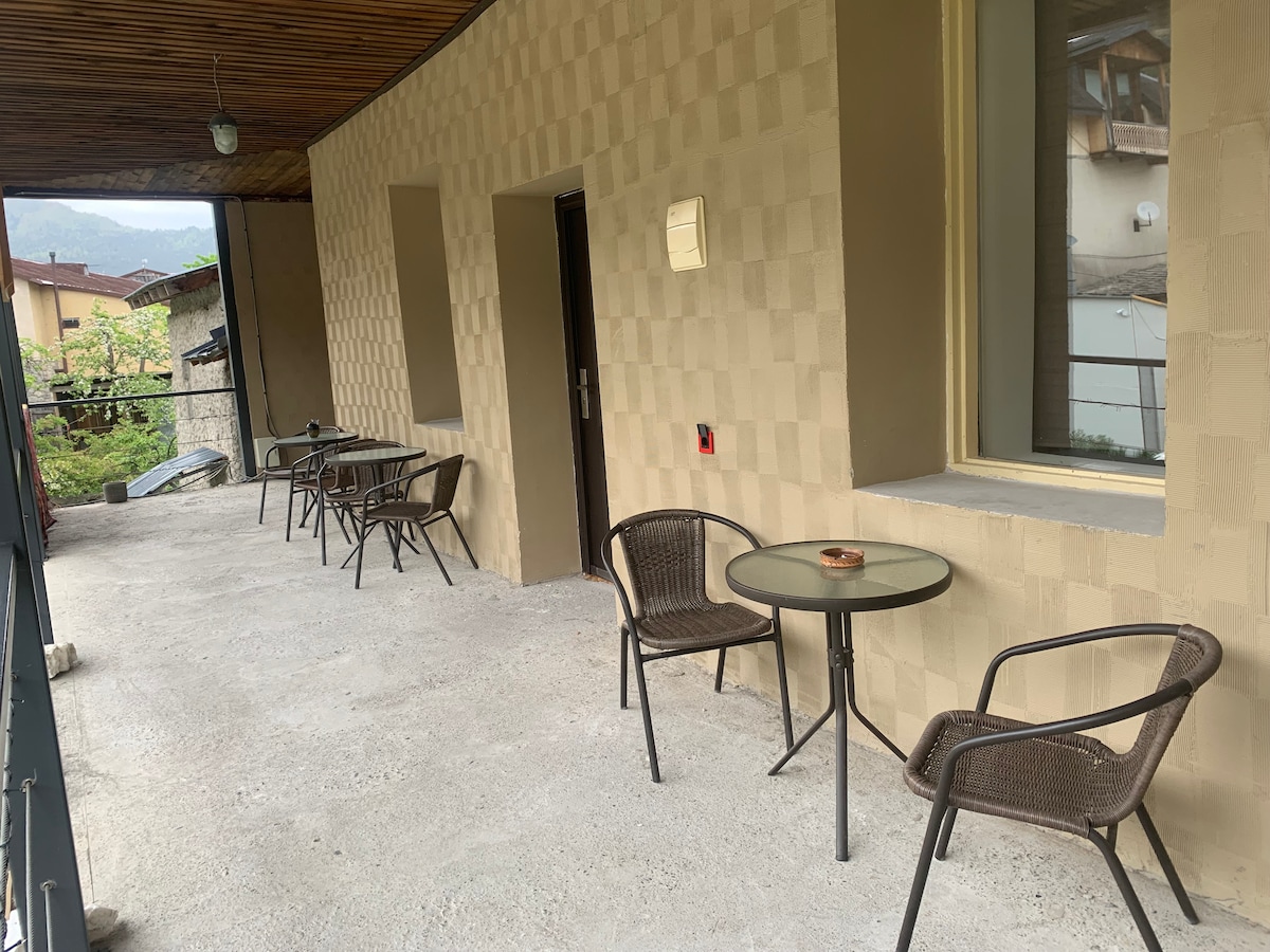 A covered outdoor terrace is displayed, featuring several small tables accompanied by chairs. The smooth concrete flooring extends along the space, with light streaming in through large windows. A peaceful view of greenery can be seen in the distance.