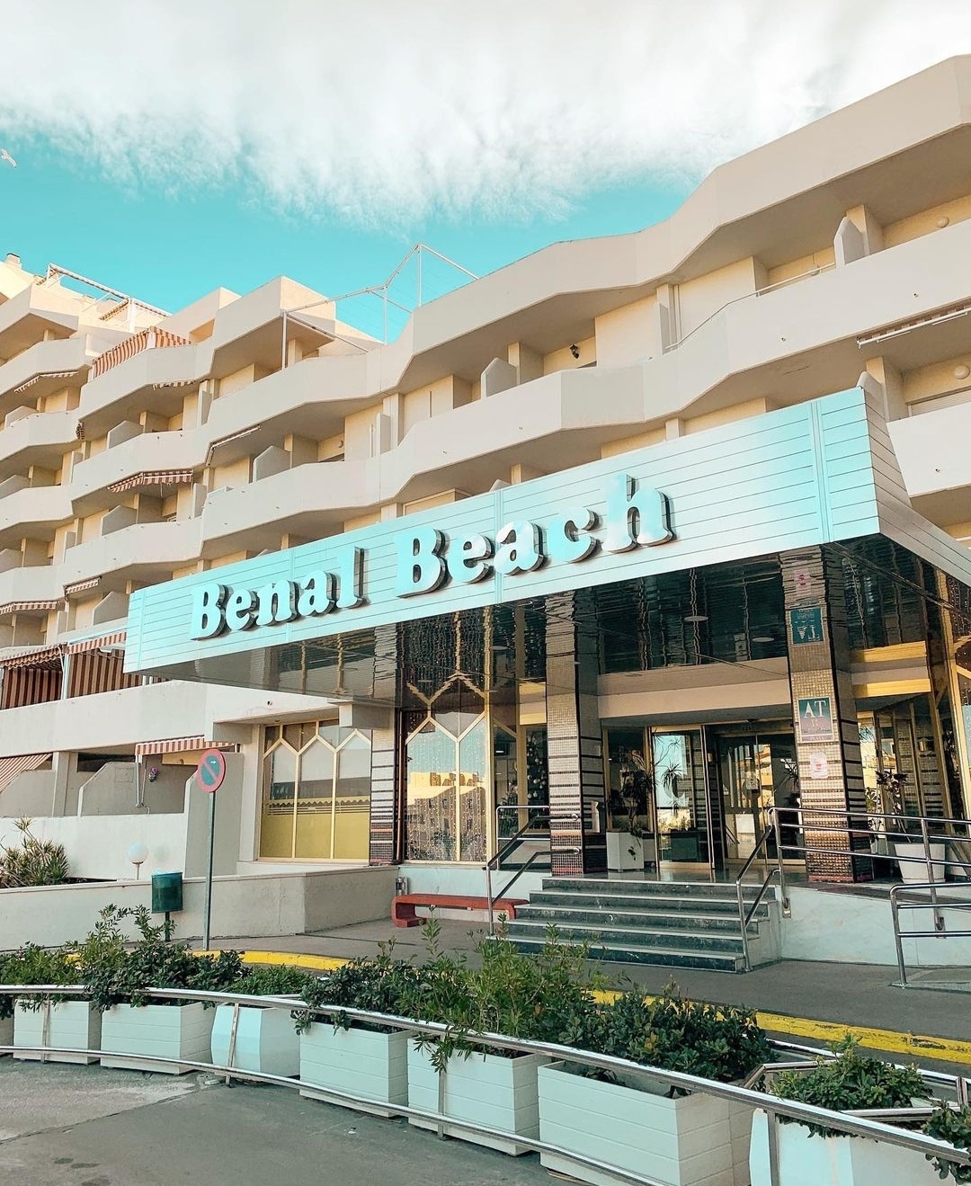 The entrance of Benal Beach is captured, showcasing the modern architecture of the building with a light blue and white facade. A landscaped area with greenery is visible in front, enhancing the welcoming appearance of the complex.