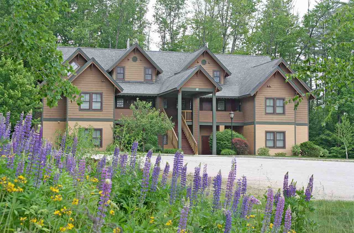 A multi-story lodge is shown, featuring a combination of wood and brown exterior. Surrounded by lush greenery, the entrance is framed by colorful wildflowers, including purple lupins and yellow blooms, creating a serene outdoor setting.
