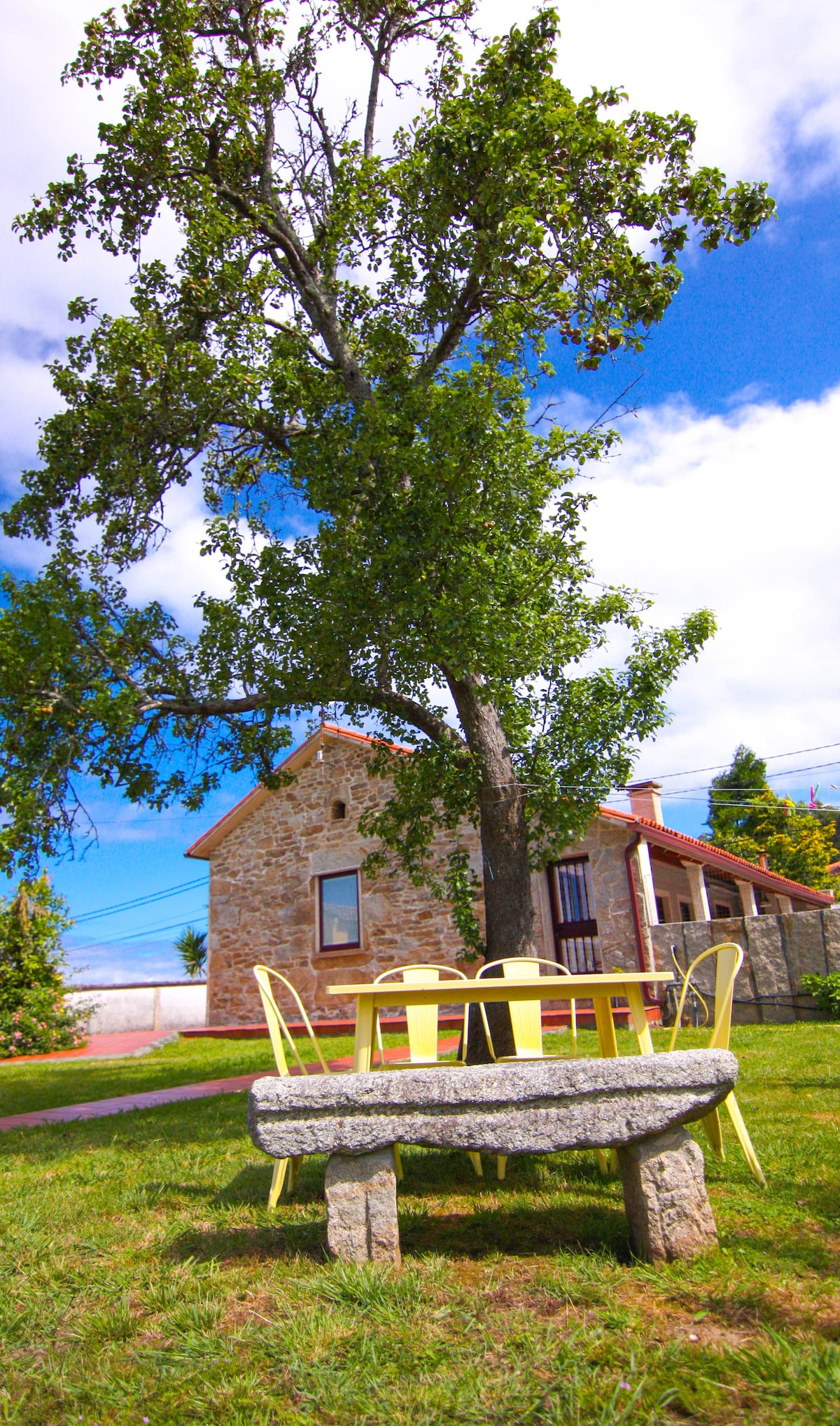 A rustic stone house is visible in the background, surrounded by greenery. A large tree provides shade over a picnic-style table with yellow chairs set on a grassy area, inviting outdoor dining or relaxation.