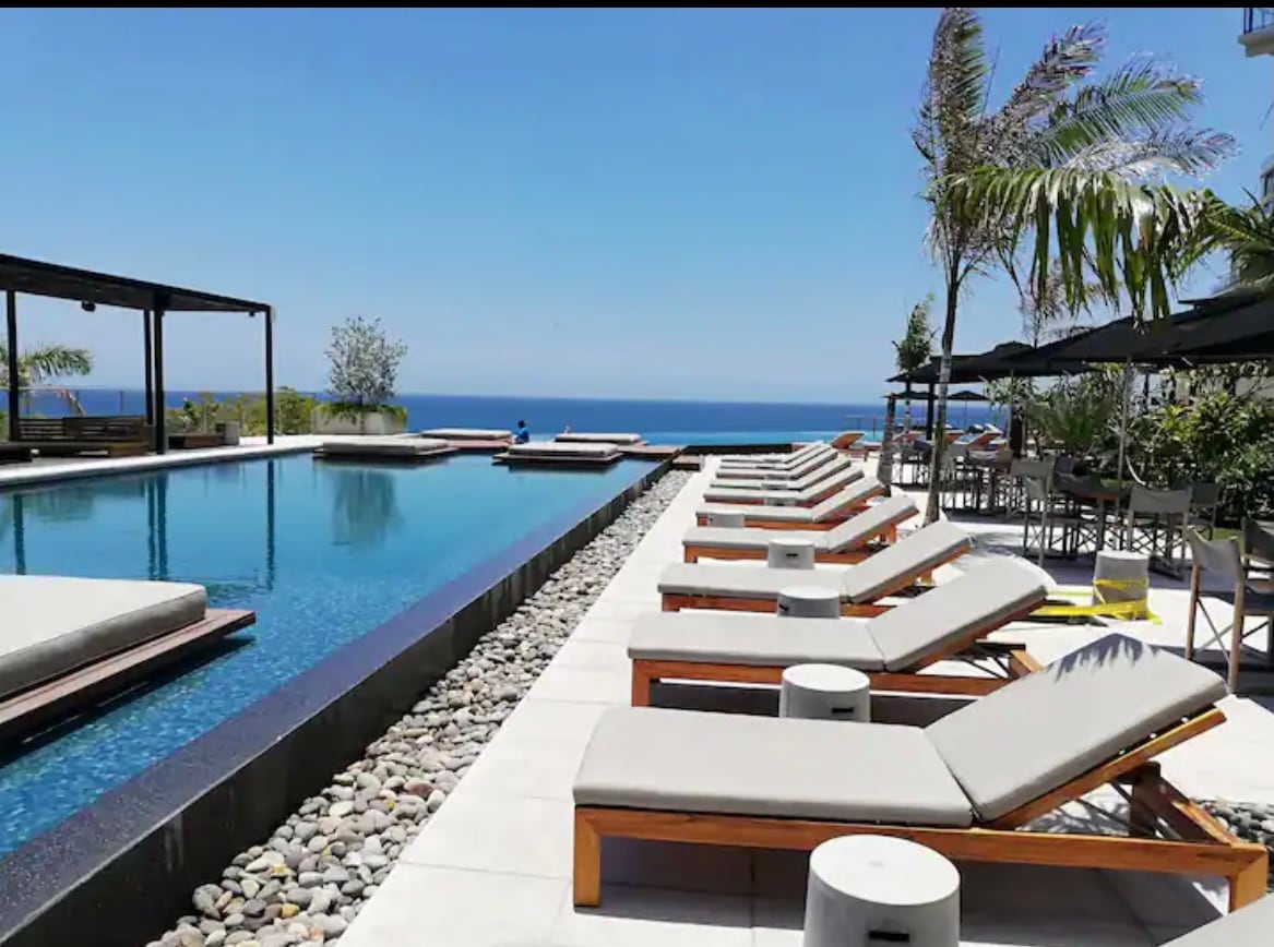 An infinity pool stretches toward the ocean, framed by wooden lounge chairs arranged neatly along the edge. Small palm trees provide dappled shade, while a seating area with tables and umbrellas is visible in the background. The clear sky enhances the serene atmosphere.