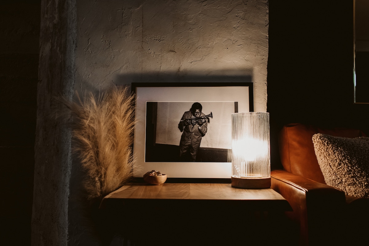 A cozy corner showcases a framed black-and-white photograph of a musician holding a trumpet. A textured lamp casts a warm glow, enhancing the space. Soft, neutral tones are present in the wall and furnishings, with decorative grasses adding a natural element.