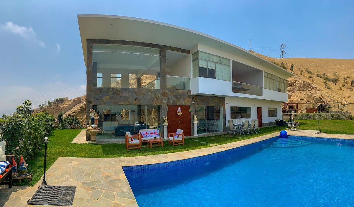 A modern two-story house is depicted with large glass walls allowing natural light. A spacious pool in vibrant blue is situated in the foreground, with ample outdoor seating areas and lush green landscaping enhancing the serene atmosphere. The surrounding hills provide a scenic backdrop.