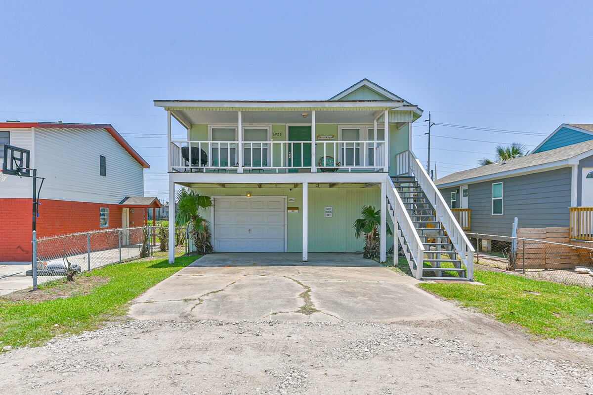 A spacious beach house stands elevated with a covered front deck. A set of stairs leads up to the main entrance. The driveway provides ample parking space, with well-maintained grass surrounding the yard. Nearby structures complete the charming neighborhood view.