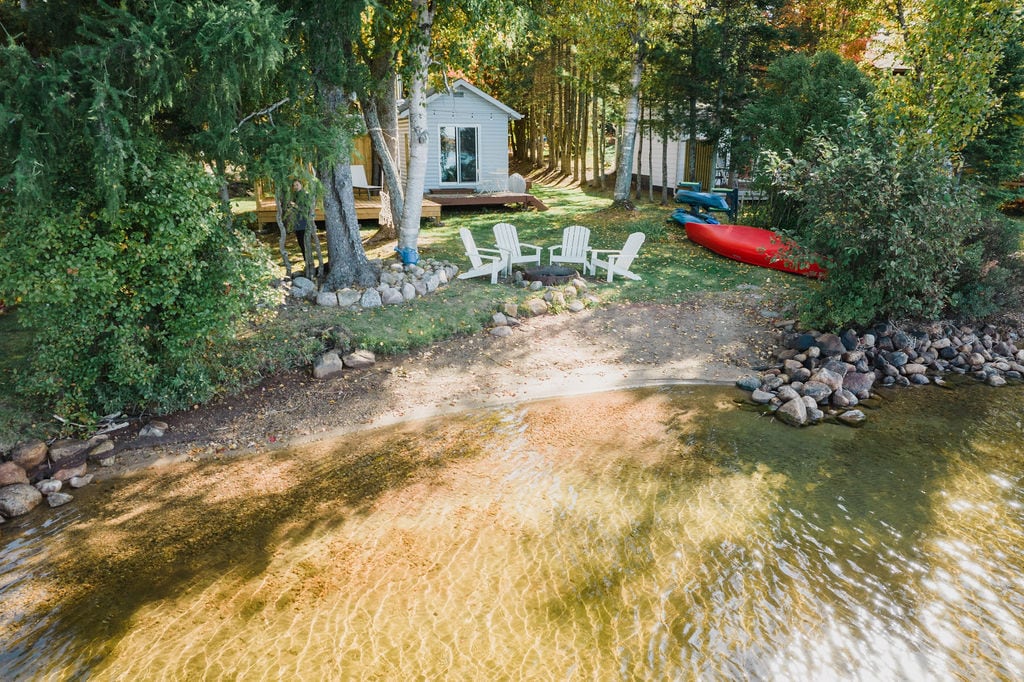A view of the cottage property from above, featuring a sandy beach area along Lake Bernard. Several white adirondack chairs surround a fire pit, with kayaks visible on the shore. Lush trees provide a serene backdrop, enhancing the cozy outdoor space.