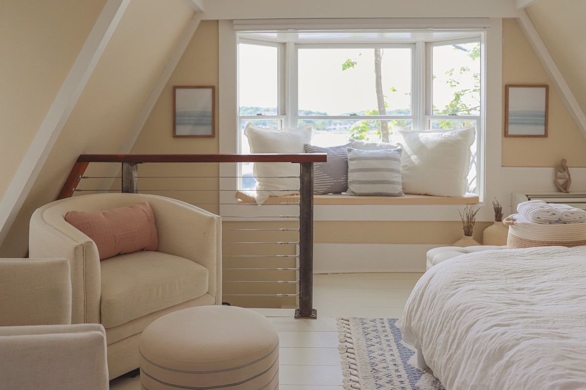 A cozy corner seating area features a light-colored chair and a round ottoman, positioned near a large window. Plush cushions rest on a built-in bench, offering a view of the surroundings. Soft, neutral tones create a calm and inviting ambiance within the space.