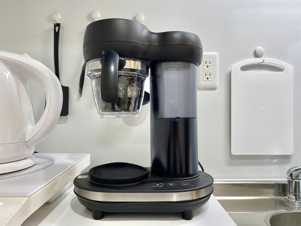 An all-in-one coffee maker is displayed on a kitchen countertop, featuring a glass carafe and a digital interface. Surrounding appliances include a kettle on the left and storage space on the right. The setup offers a modern and functional design for coffee preparation.
