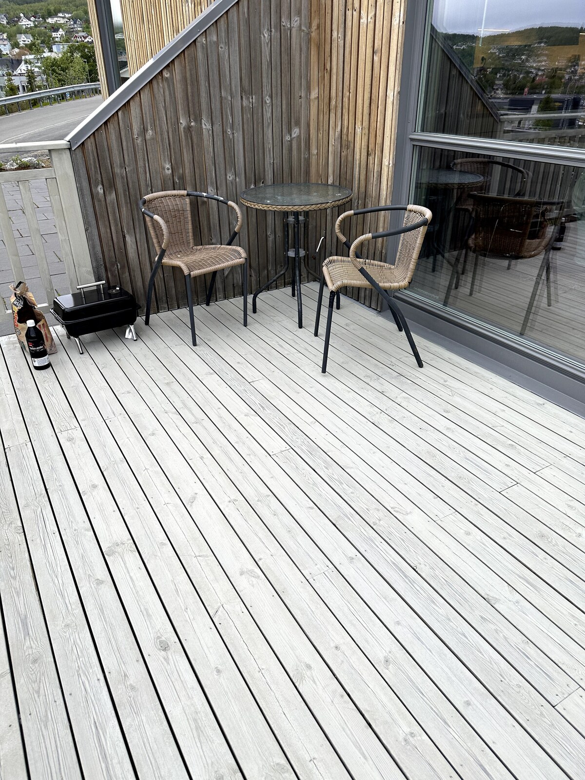 A balcony area is equipped with a small round table and two woven chairs. The wooden decking features a light-colored finish, and a cooler is positioned alongside the table. Glass doors provide access to the interior space.