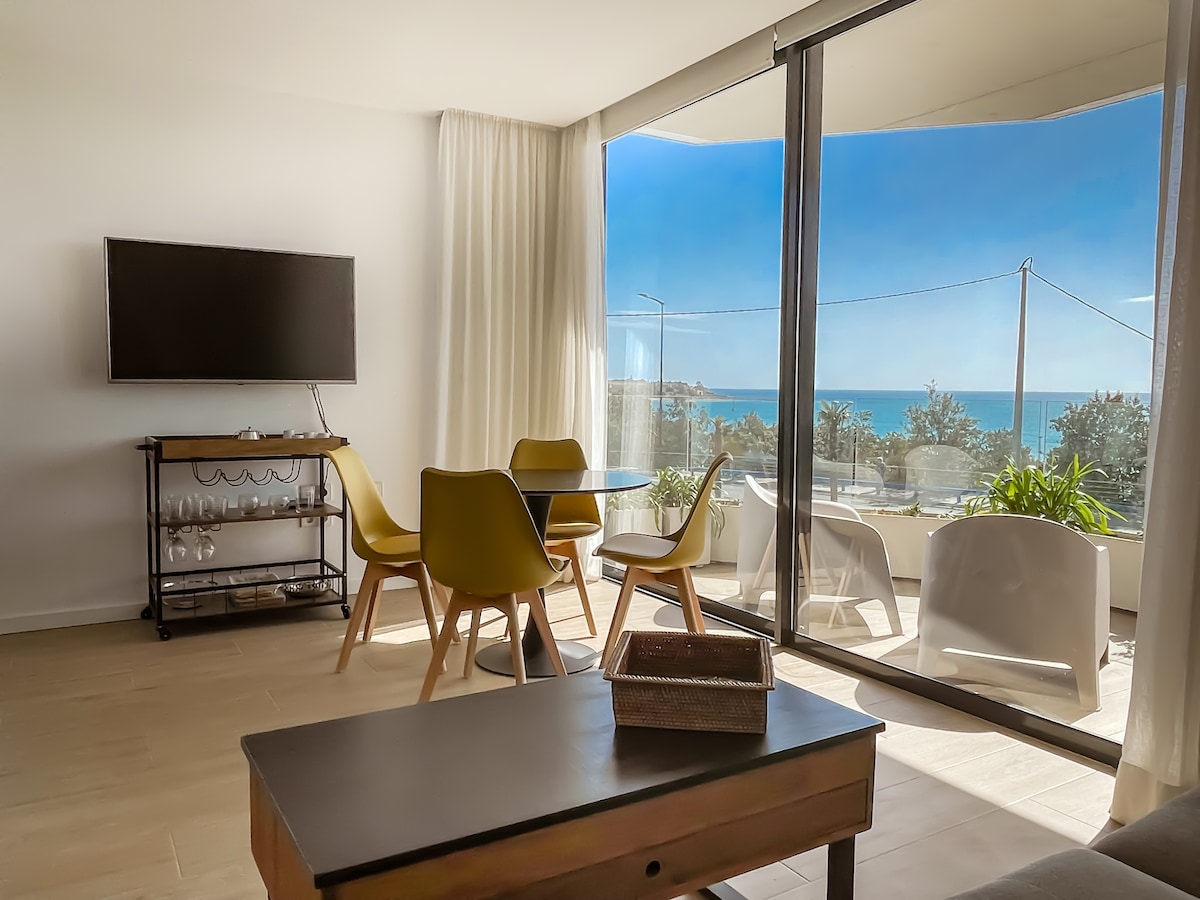 A modern living area features a glass wall that opens to a stunning view of the sea. A stylish dining table is surrounded by yellow chairs, while a sleek television is mounted on the wall. Natural light fills the space, complemented by light drapes.