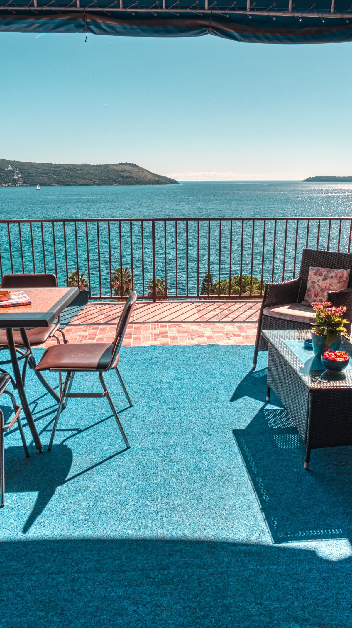 An outdoor terrace offers views of the sea, furnished with a table and chairs. A small coffee table displays a bowl of fresh fruit and a vase with flowers. The flooring is a vibrant blue, contrasting with the natural colors of the surrounding landscape.