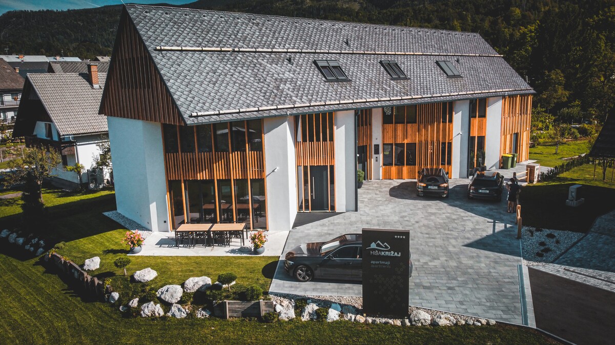 The modern building features a combination of wooden and white finishes, with large windows allowing natural light to fill the space. A paved driveway leads to the entrance, where vehicles are parked. Well-maintained landscaping with stones and greenery surrounds the property.