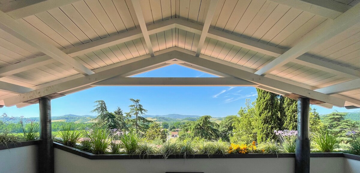 A scenic view is framed by an angled ceiling with exposed beams. Lush greenery and trees are visible outside, extending into the rolling hills of the Tuscan countryside under a clear blue sky.