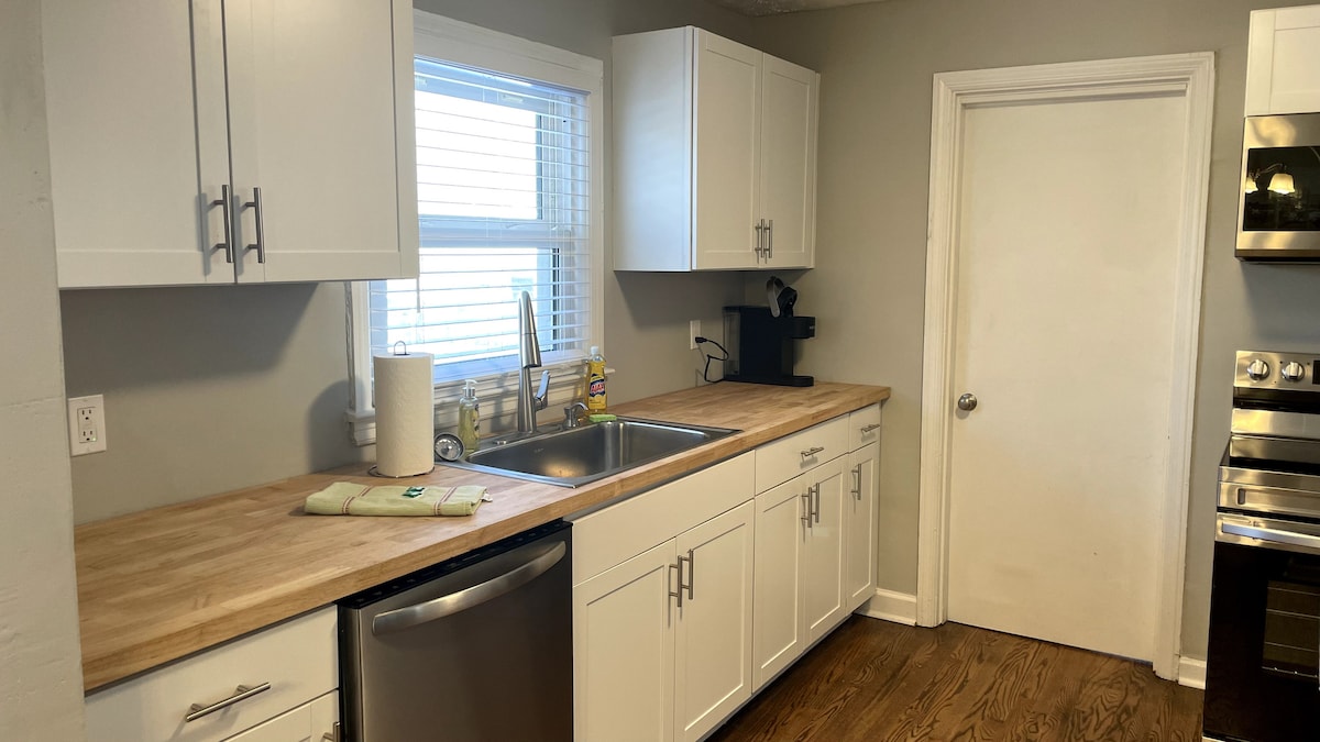 A well-equipped kitchen features white cabinetry and a wooden countertop. A stainless steel dishwasher is positioned beneath the sink, which is situated between two windows providing natural light. A coffee maker is placed on the countertop, complementing the minimalist design.