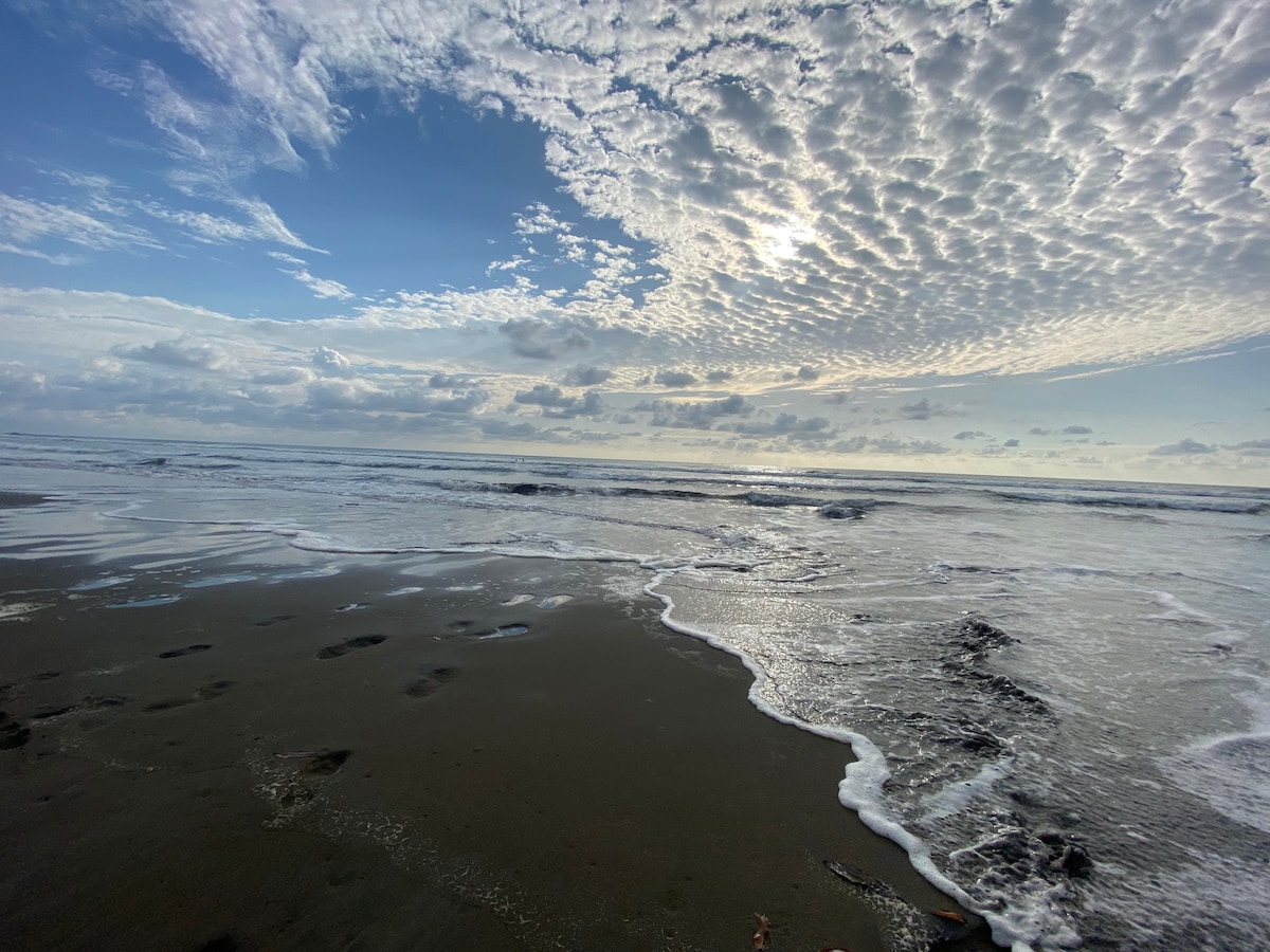 A wide view of a beach presents gentle waves lapping the shore, with a sandy surface marked by footprints. The sky is filled with textured clouds, reflecting light that creates a serene atmosphere over the ocean.
