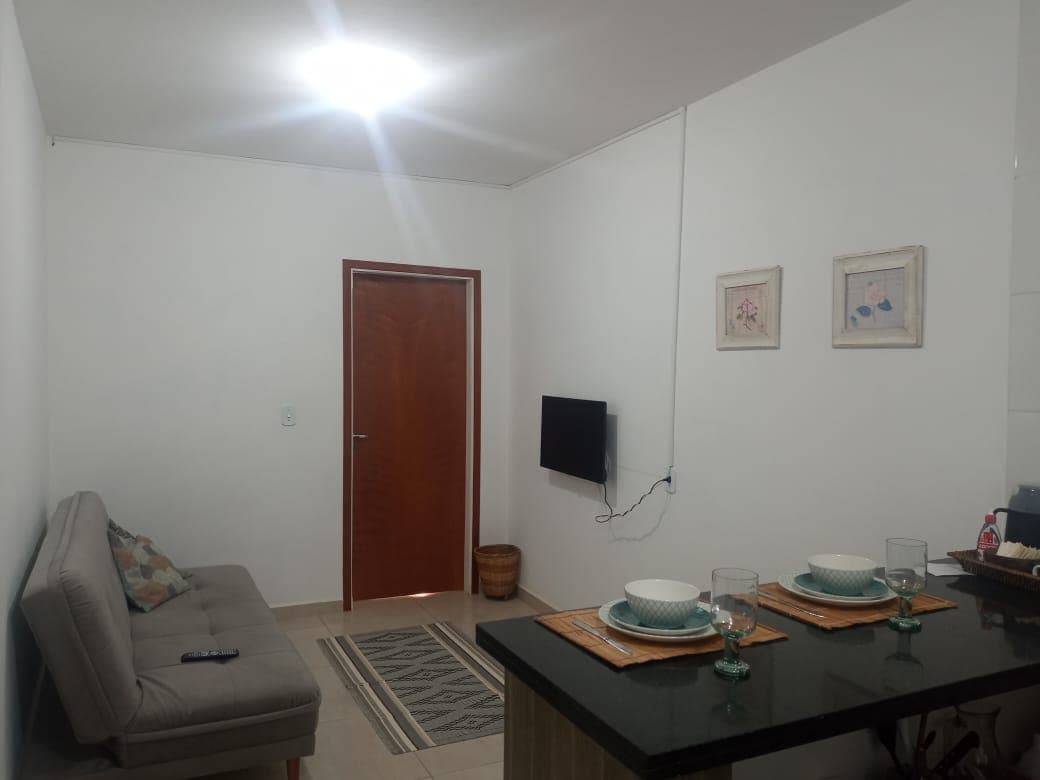 A bright living area features a colorful sofa bed along one wall, complemented by a coffee table set with dishes and glasses. A wall-mounted TV is positioned across from the seating. The space is illuminated by a ceiling light, creating a welcoming environment.