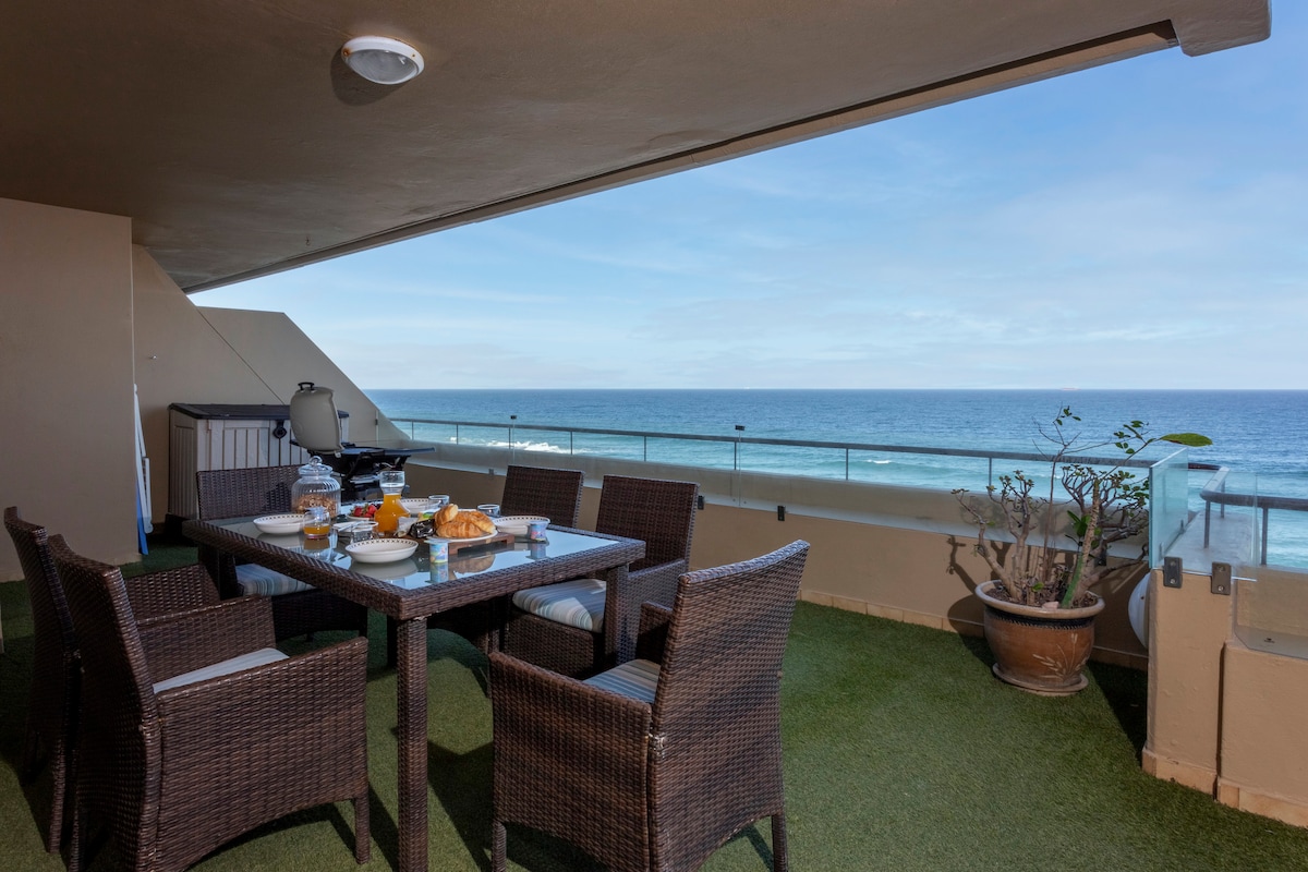 A spacious patio offers a panoramic view of the Indian Ocean, featuring a dining table surrounded by chairs. A gas braai is positioned nearby, set against a backdrop of lush greenery and the sea beyond, creating an inviting outdoor space for gatherings.