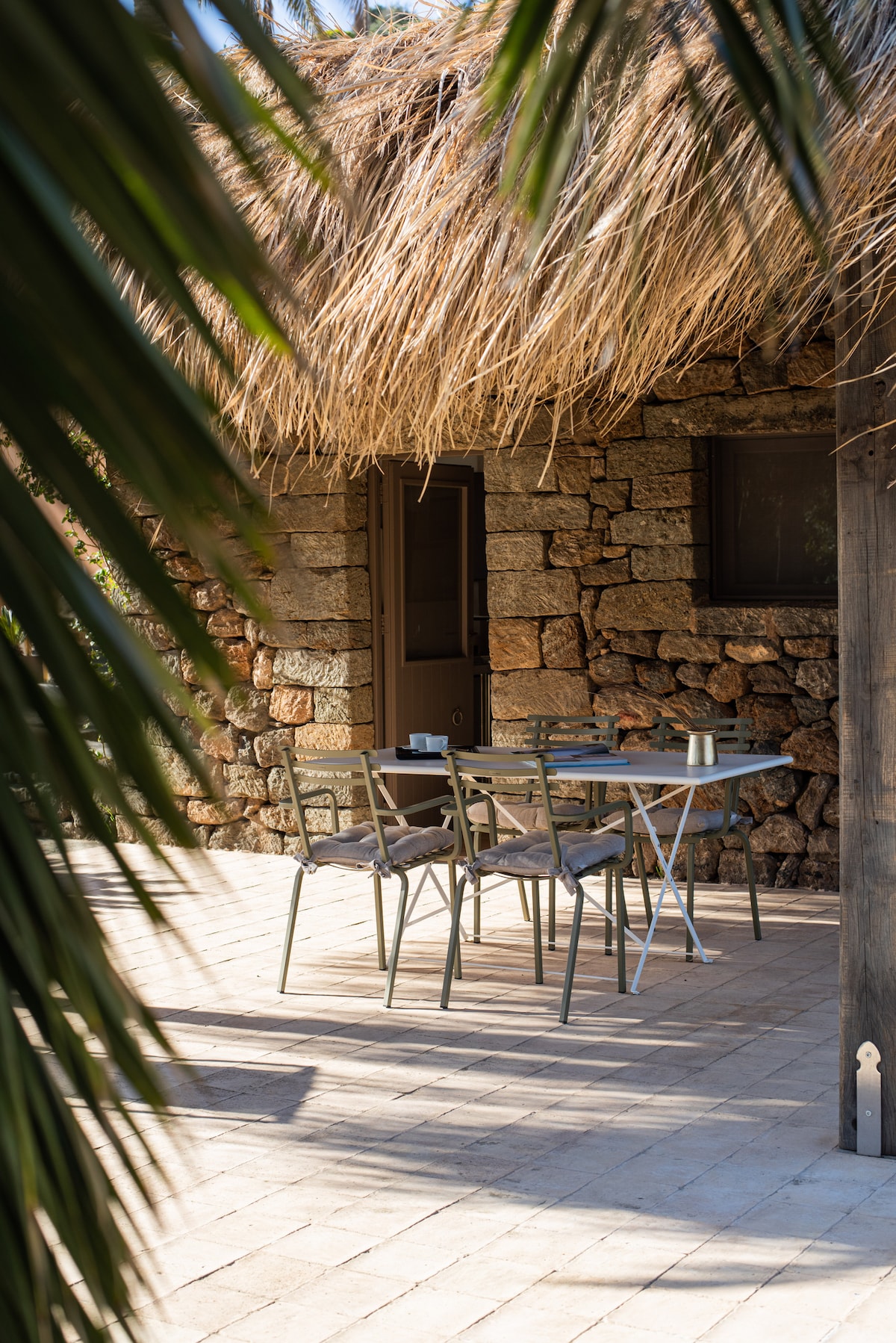 An outdoor dining area is visible, featuring a table with chairs positioned under a thatched roof. Natural stone walls and windows are partially obscured by surrounding greenery, creating a serene and private space for relaxation.