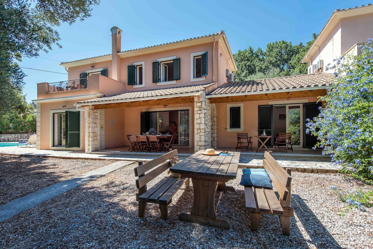 The villa features a spacious outdoor area, highlighting a wooden dining table with benches surrounded by gravel. The bright facade showcases two levels, with large glass doors that provide access to the patio and views of the pool nearby. Lush greenery is visible in the background.