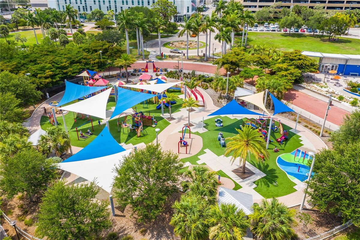 A vibrant playground is situated in a spacious park, featuring colorful fabric canopies providing shade. Various play structures are visible, surrounded by lush greenery and palm trees. Well-maintained grass areas and pathways are evident, inviting families to enjoy outdoor activities.