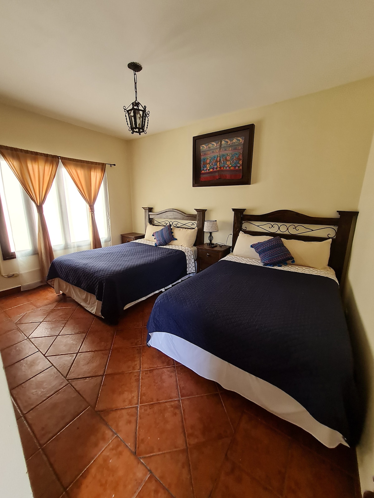 The bedroom features two spacious beds with dark blue bedding, positioned opposite each other. Light streams through two large windows, adorned with flowing brown curtains. Wooden nightstands accompany each bed, providing a cozy and functional space.