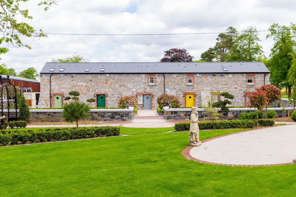 A charming stone building is set against a lush green lawn, featuring colorful doors that add a distinctive touch. Neatly trimmed bushes and small trees create a serene garden atmosphere, enhancing the peaceful surroundings.