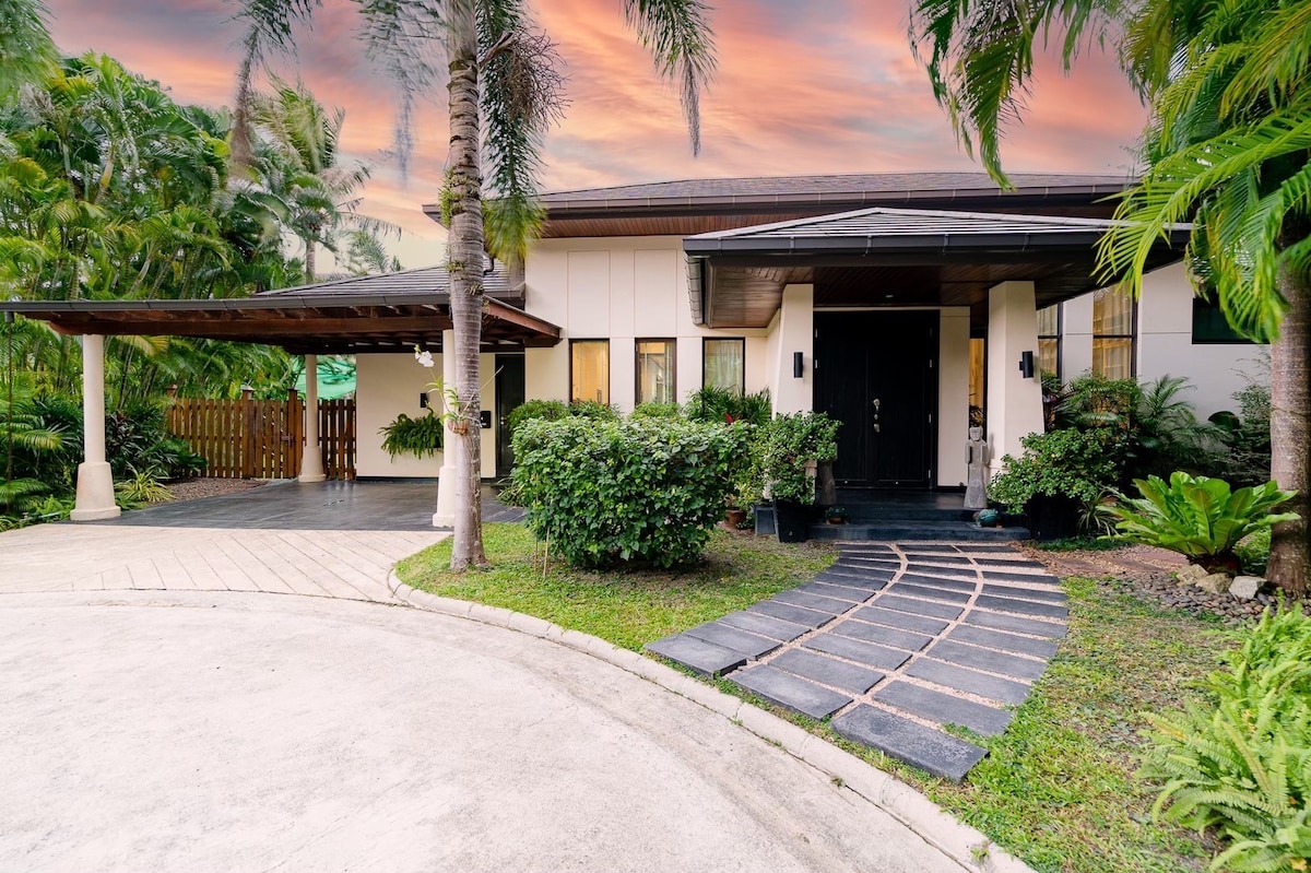 A modern exterior is presented, featuring a welcoming entrance adorned with lush greenery. The driveway is crafted from smooth stones, leading to a covered entryway. Large windows provide glimpses into the interior, while palm trees frame the scene against a colorful sky.