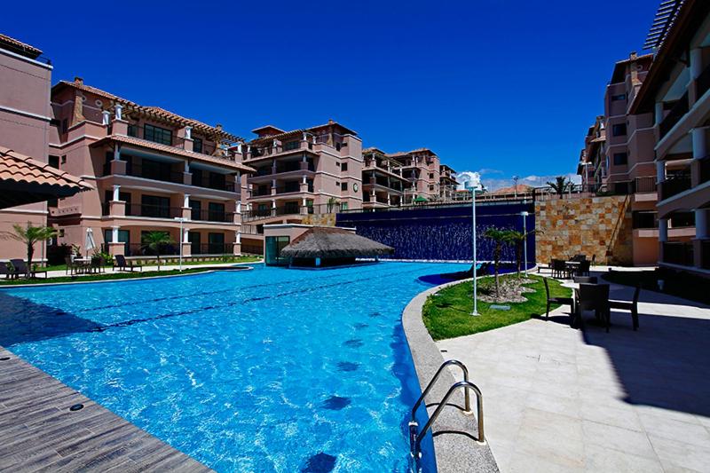 A large outdoor pool is surrounded by landscaped areas and lounge chairs. The pool area is complemented by a waterfall feature, and several multi-story buildings provide a scenic backdrop. The clear blue sky enhances the inviting atmosphere of the space.
