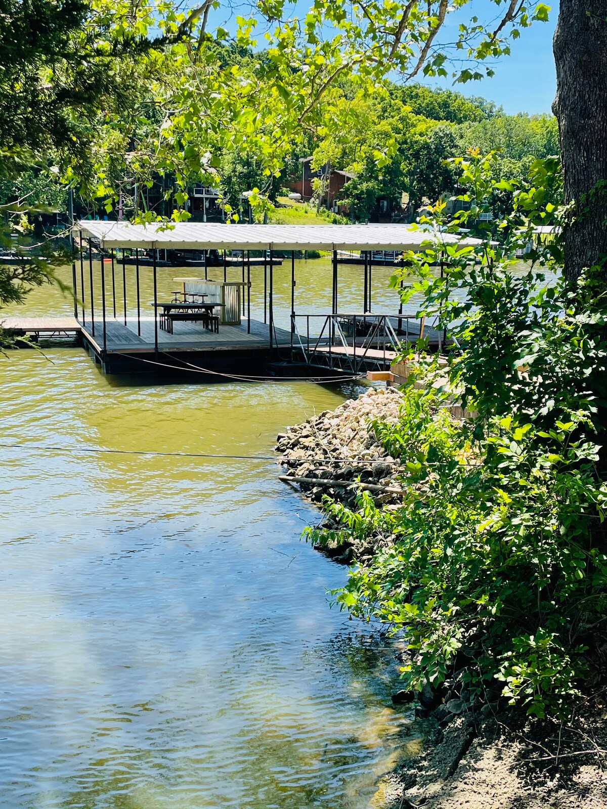 A private dock extends over the calm water, featuring a shaded area suitable for dining. Lush green trees frame the scene, and the shoreline reveals a rocky edge, inviting opportunities for fishing or swimming.