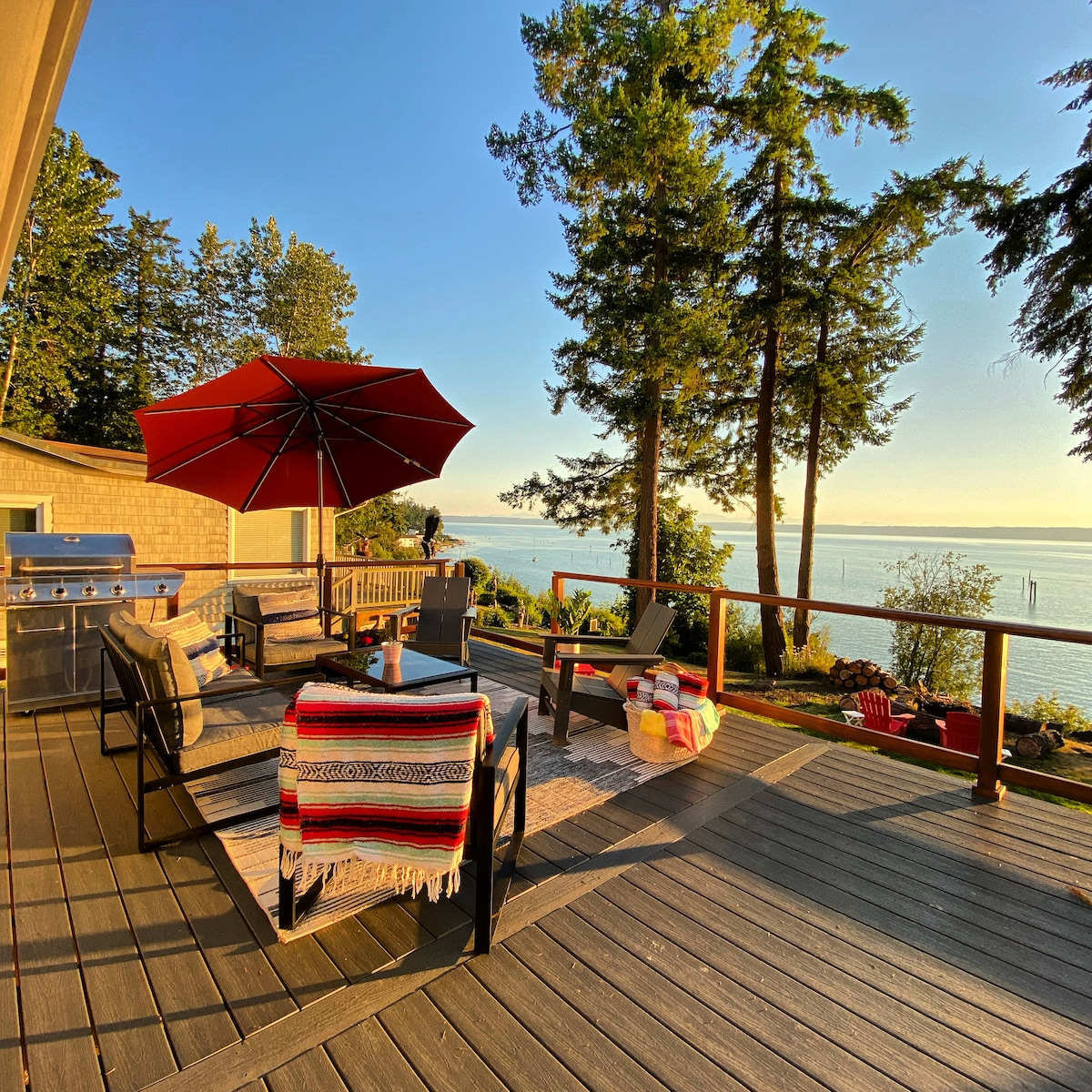 A spacious deck is presented with comfortable seating arrangements, including lounge chairs and a table shaded by a large umbrella. The surrounding tall trees provide a natural frame while scenic views of the water and mountains are visible in the background.
