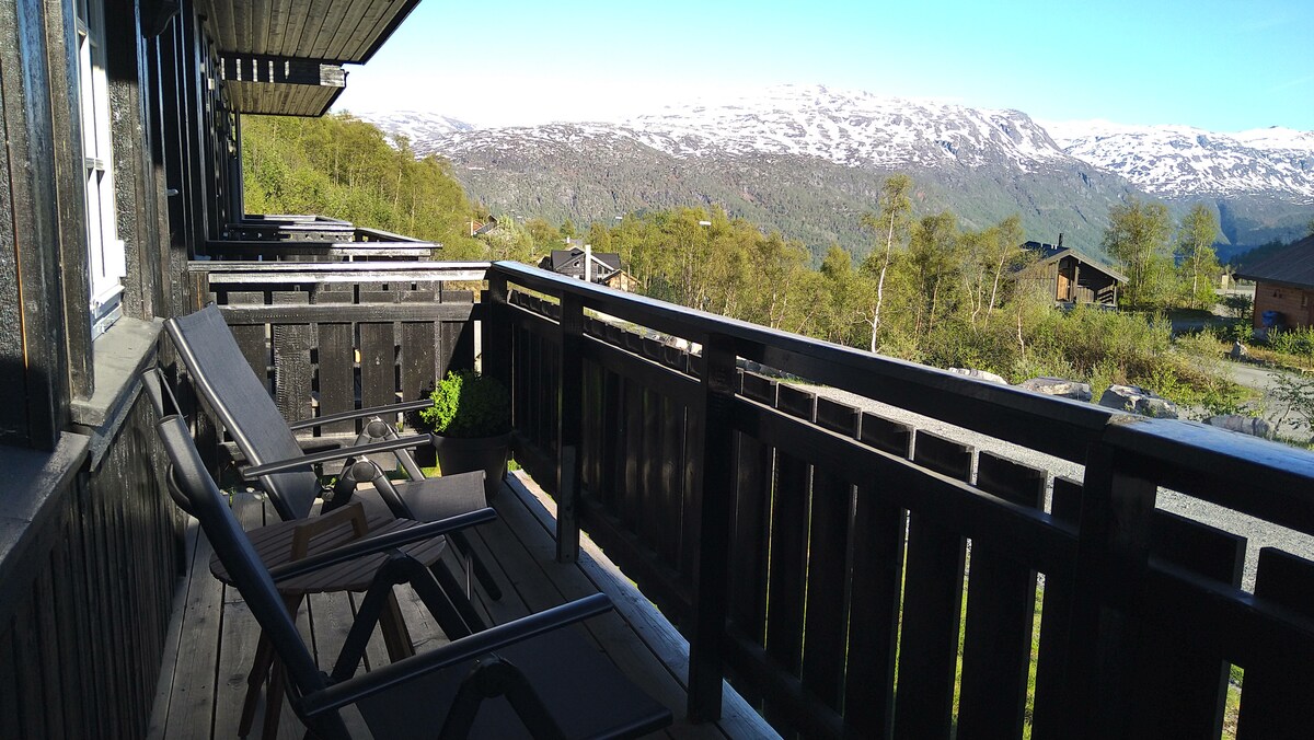 A balcony with wooden railings overlooks a valley filled with greenery and distant snow-capped mountains. Two lounge chairs are positioned for relaxation, enhancing the outdoor seating space. The bright blue sky completes the scenic view, inviting tranquil moments in nature.