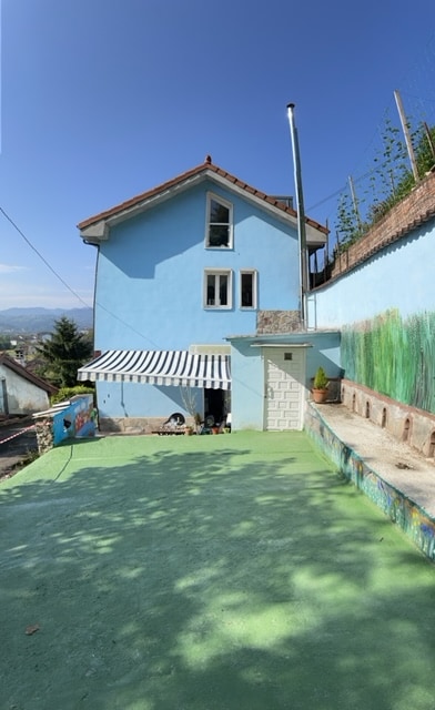A two-story house with a light blue exterior is pictured, featuring multiple windows and a striped awning. A green outdoor area extends in front of the building, coupled with a textured wall showcasing a nature scene. The space has a clear view of the surroundings.