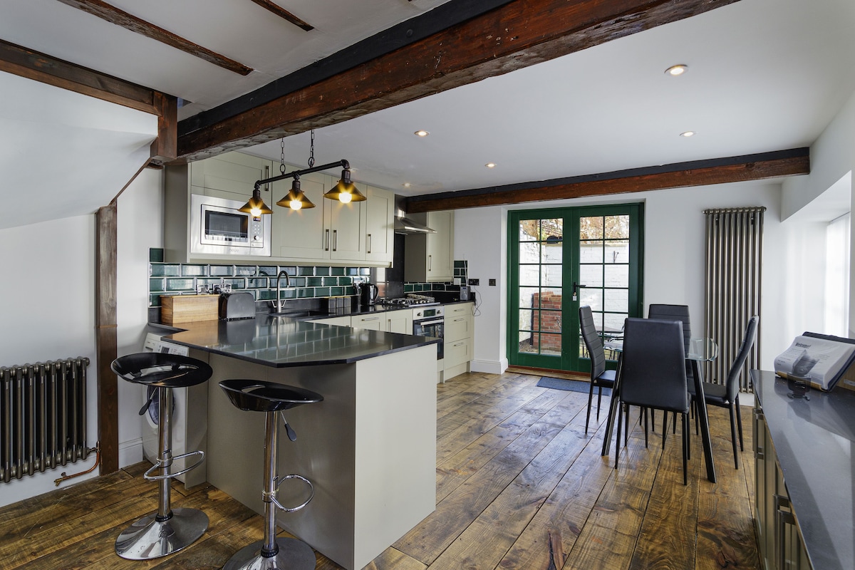 An open-plan living area features a stylish kitchen with modern appliances and a spacious island. A wooden flooring adds warmth, while tall windows with green frames allow natural light to brighten the space. Seating is provided for six around a sleek dining table.