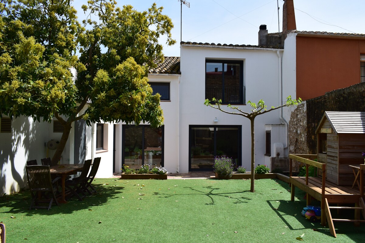 A spacious garden area features green grass and shaded seating, with a large tree providing natural cover. The house is visible in the background, showcasing expansive windows that enhance natural light. A play structure is present, adding a recreational element for children.