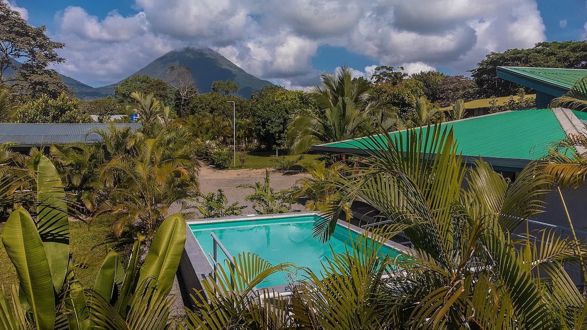 A private swimming pool is framed by lush tropical greenery, with views of the Arenal Volcano in the background. The clear blue water reflects the sky, while the surrounding landscape showcases a variety of vibrant plants and trees under partly cloudy skies.